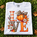 a light tan t-shirt featuring a large autumn-themed graphic that spells 'LOVE' with a pumpkin, leopard print bow, buffalo check 'L', leopard print 'O', and orange dotted 'E', 