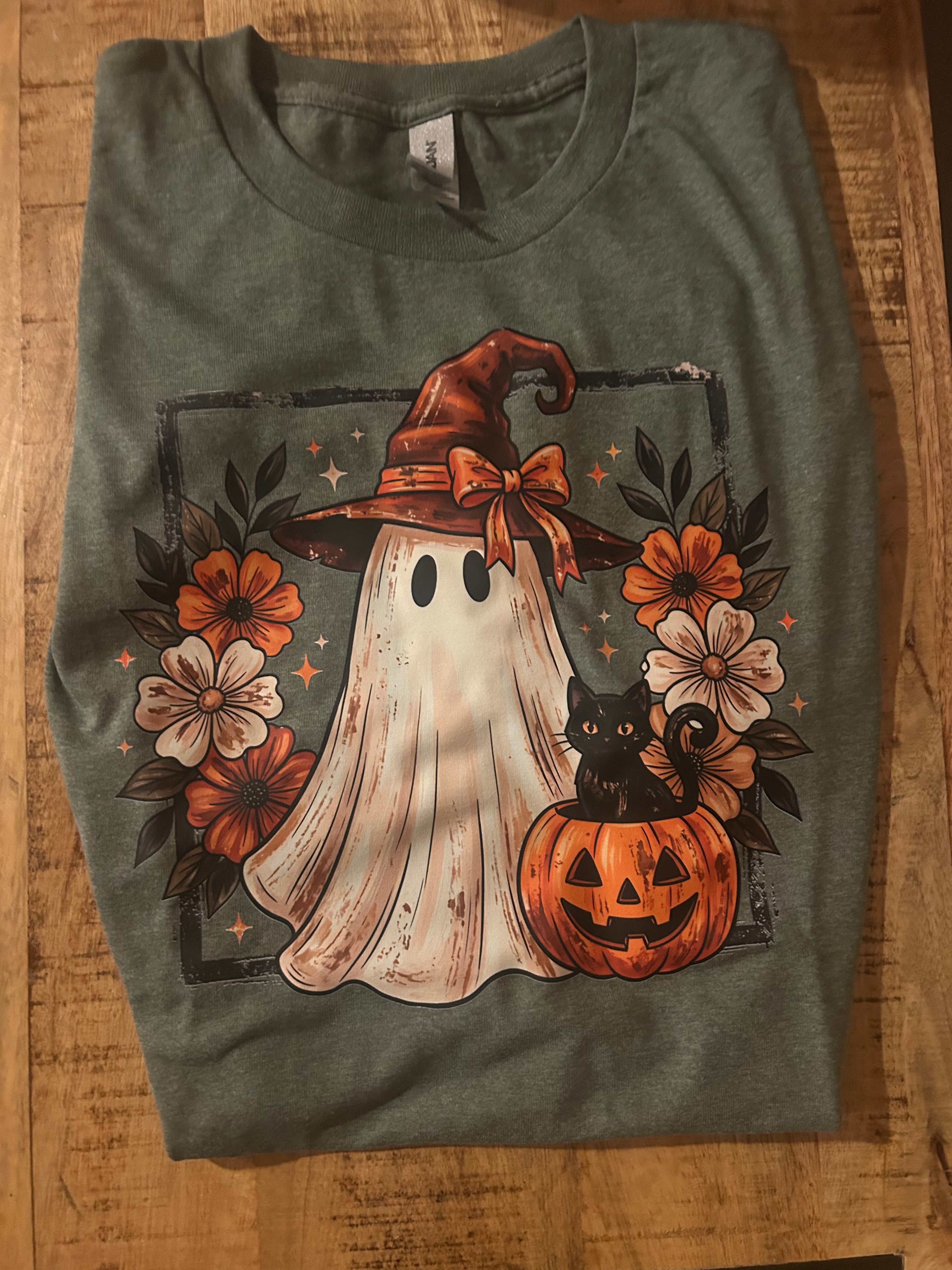  olive green t-shirt featuring a Halloween graphic print of a ghost in a witch hat, a black cat on a pumpkin, and orange flowers.